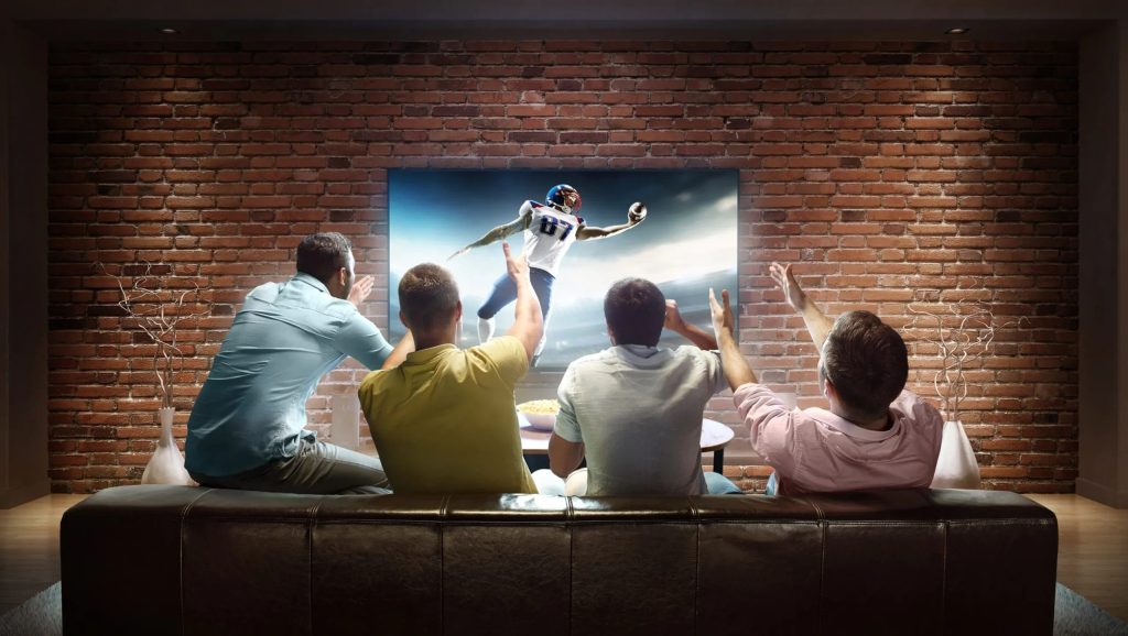 Fire mandlige venner sidder i en sofa og jubler over en amerikansk fodboldkamp på et stort tv foran en murstensvæg