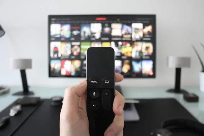 Hånd holder en Siri Remote-fjernbetjening foran et fjernsyn, der viser en streamingtjenestes brugerflade med film- og serieplakater.