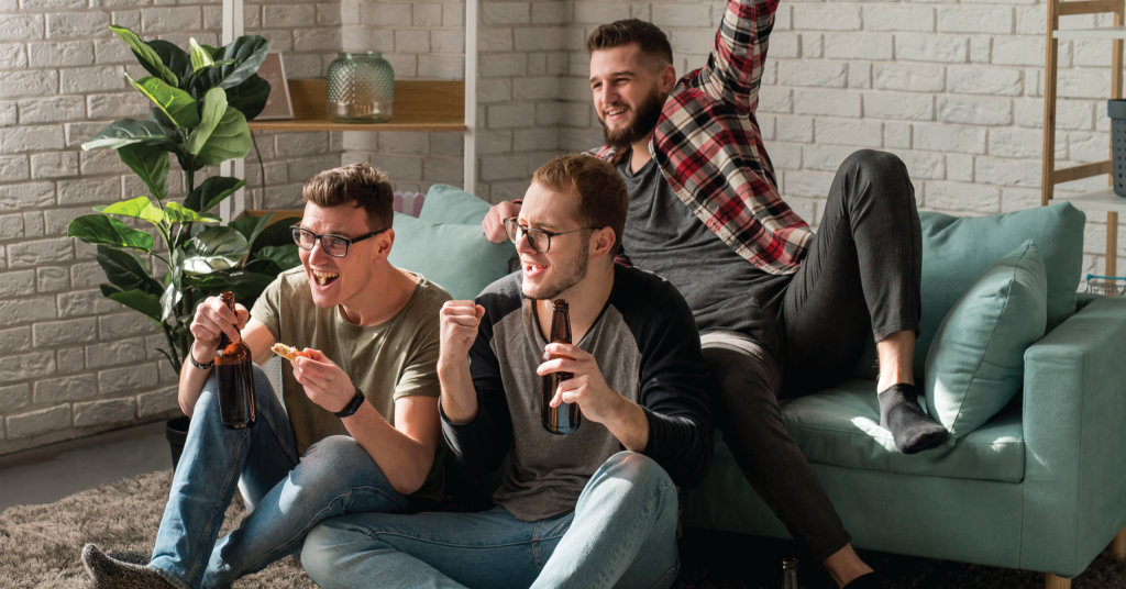 Tre glade mænd ser sport, drikker øl og jubler i en stue.